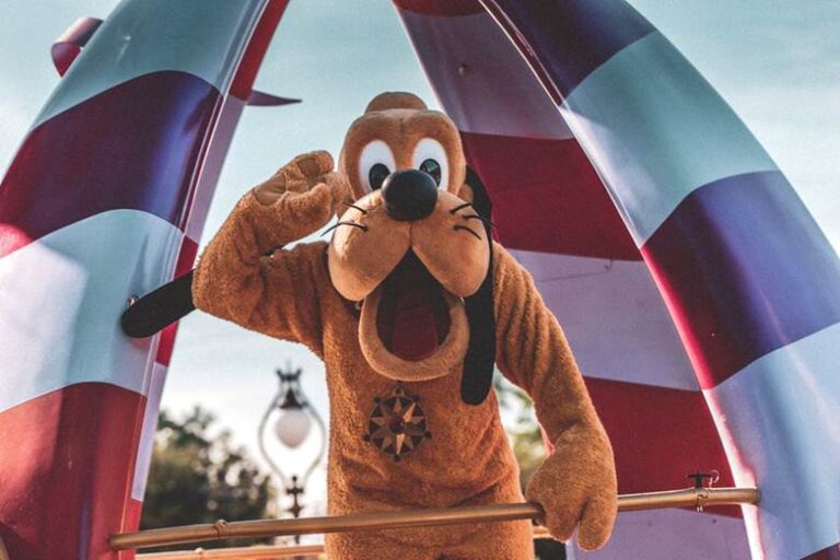 Mascot in a dog costume waving at a family-friendly amusement event with red and white decorations in the background.