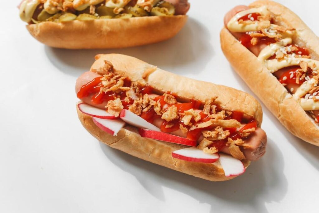 Close-up of a hot dog topped with sliced radishes, ketchup, and crispy fried onions on a white background