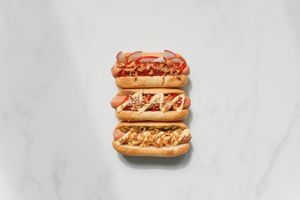 Overhead view of three hot dogs with creative toppings like radishes, fried onions, pickles, and sauces on a marble surface