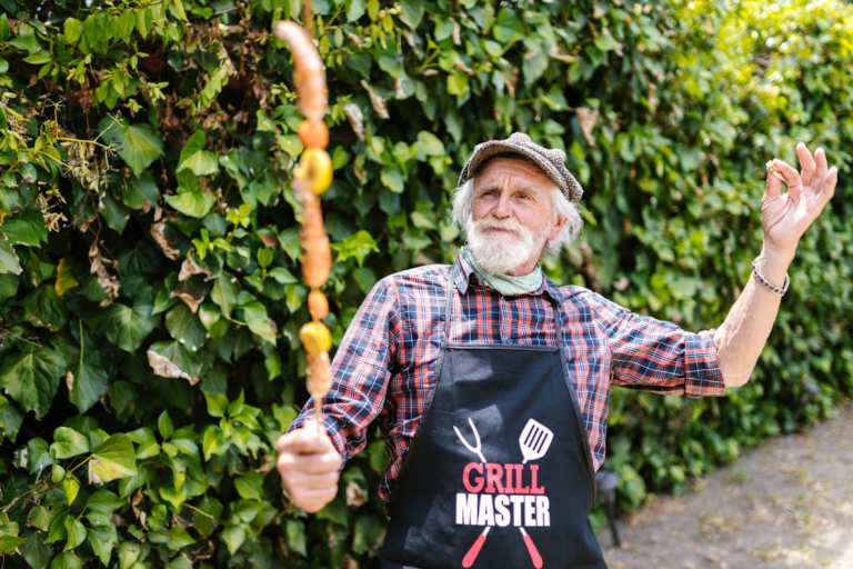 Elderly man in a “Grill Master” apron proudly holding a skewer of grilled sausages and veggies, striking a celebratory pose outdoors