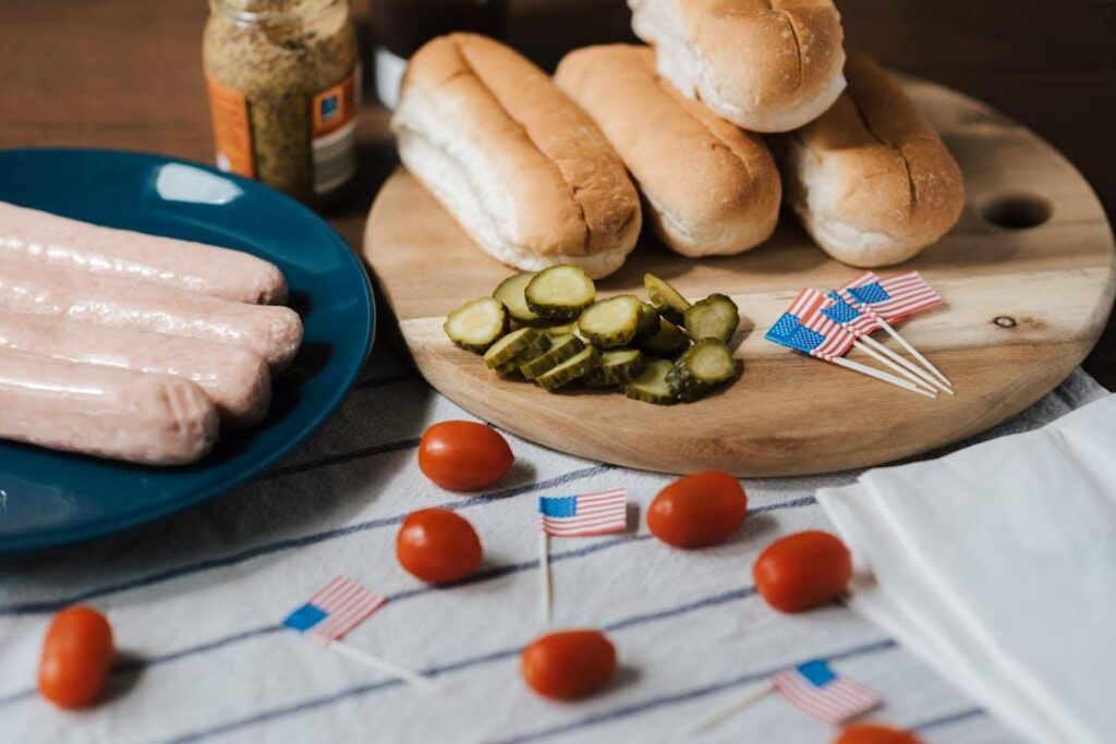 A picnic spread with hot dog buns, pickles, tomatoes, sausages, and mini American flags arranged on a wooden board and blue plate