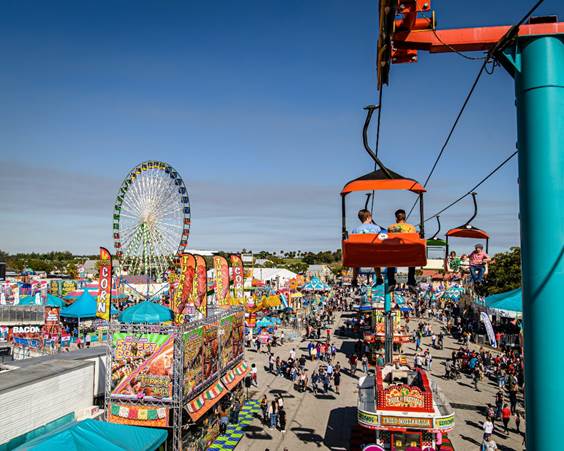 A bustling county fair with food stalls, a Ferris wheel, and a skyride carrying visitors over the crowd under a clear blue sky.