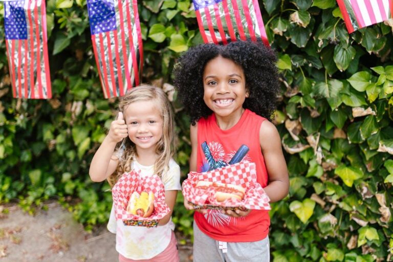 Two young children smiling and holding decorated hot dogs in paper trays, standing in front of American flags and greenery