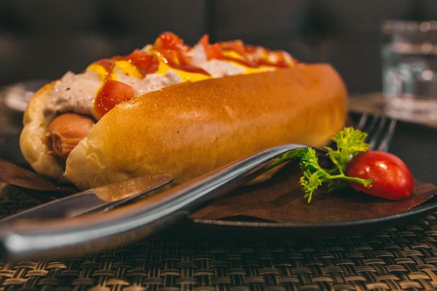 a hot dog with extra meat on top of it with ketchup and mustard, placed on a plate with a fork