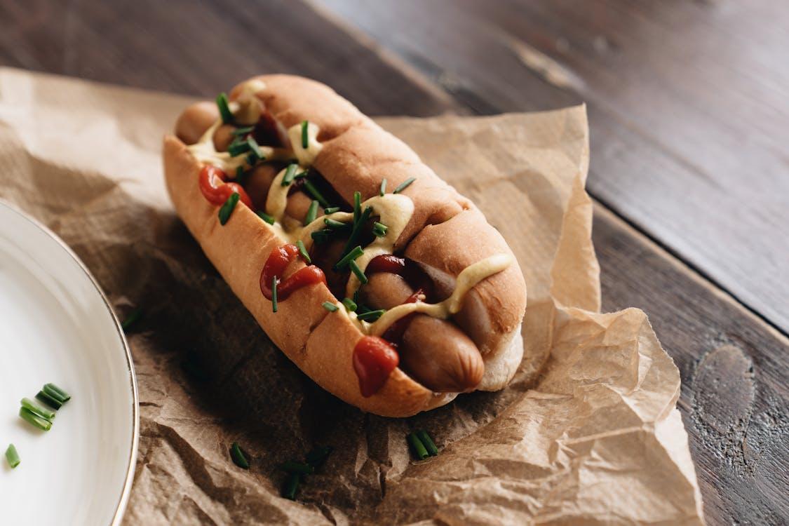 A close-up of a gourmet hot dog with mustard, ketchup, and chives on brown paper