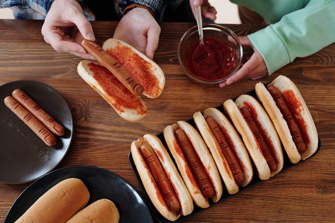 A child and adult prepare spooky Halloween-themed hot dogs with ketchup on a wooden table, arranging them neatly on a black tray.