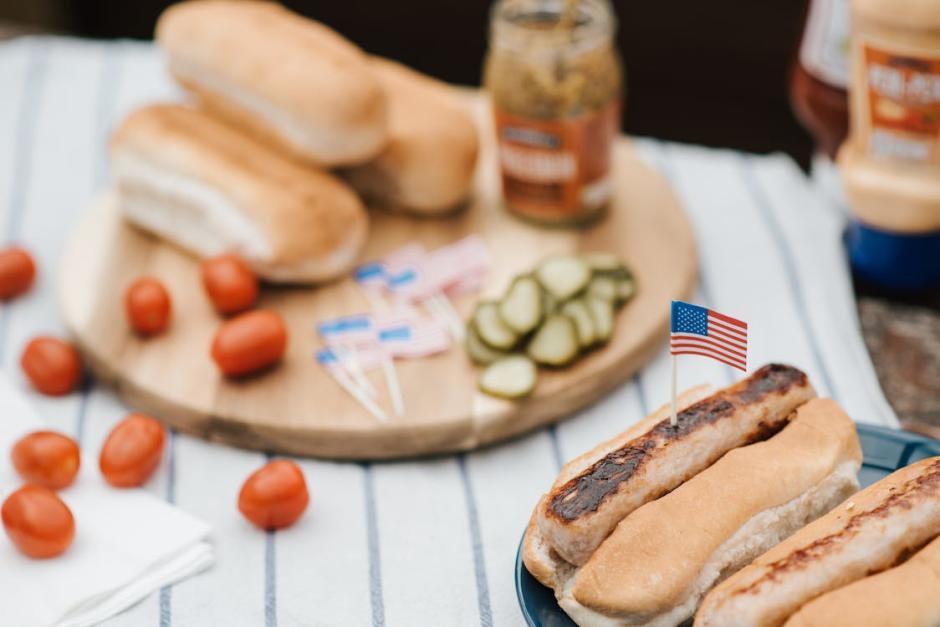 A plate of hot dogs with American flags sits in front of a spread of pickles, mustard, buns, and cherry tomatoes on a striped cloth.