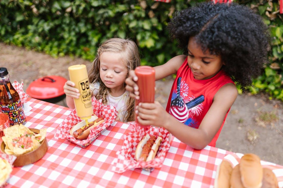 Two young girls at an outdoor picnic table decorate their hot dogs with ketchup and mustard