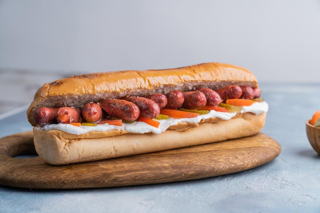 A gourmet-style hot dog topped with mini sausages, pickles, carrots, and cream on a wooden board.