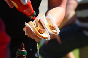 Close-up of a person topping two hot dogs with ketchup at an outdoor event