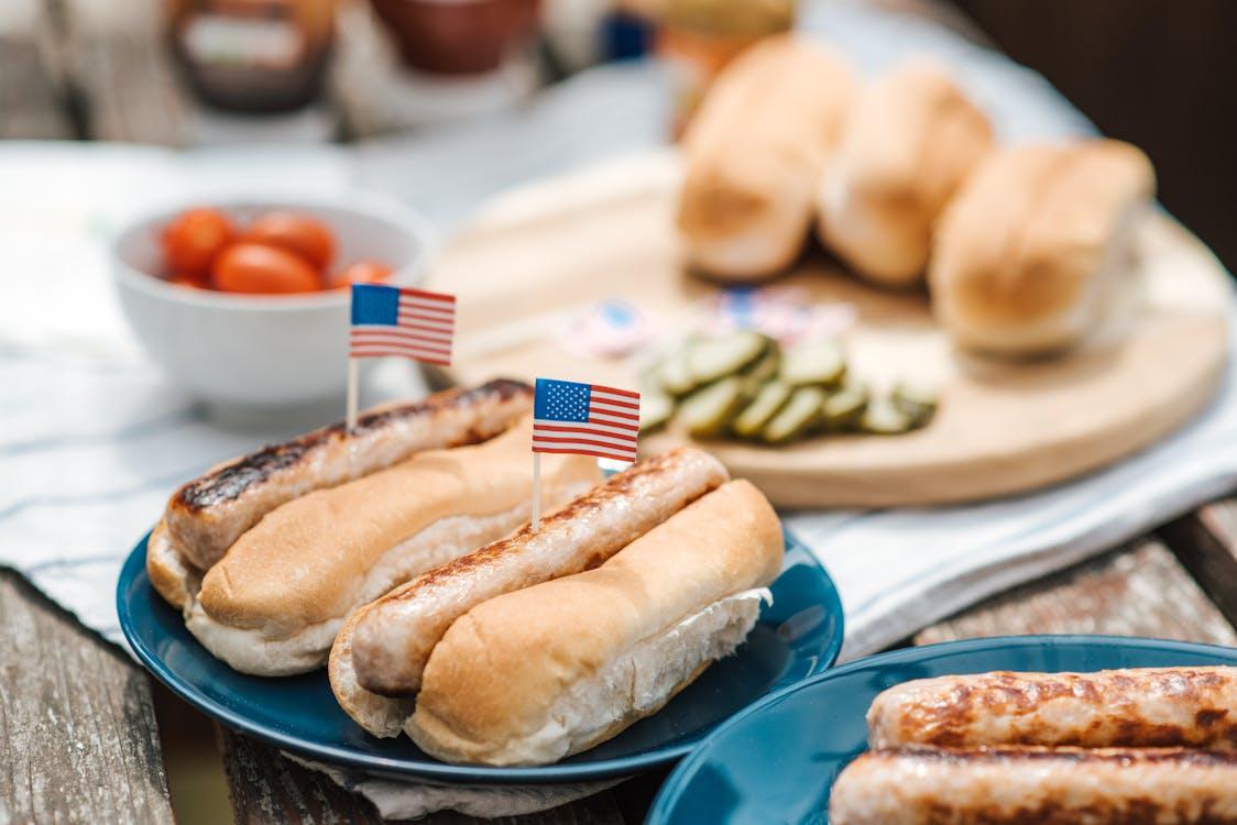 Grilled hot dogs with small American flags served on blue plates at an outdoor cookout.