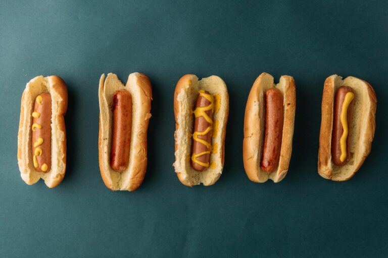 Five hot dogs with simple mustard toppings lined up against a teal background