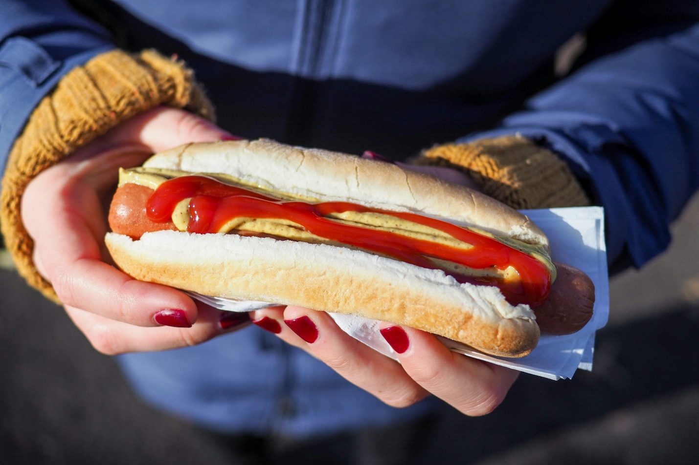 Person holding a freshly prepared hot dog with mustard and ketchup outdoors