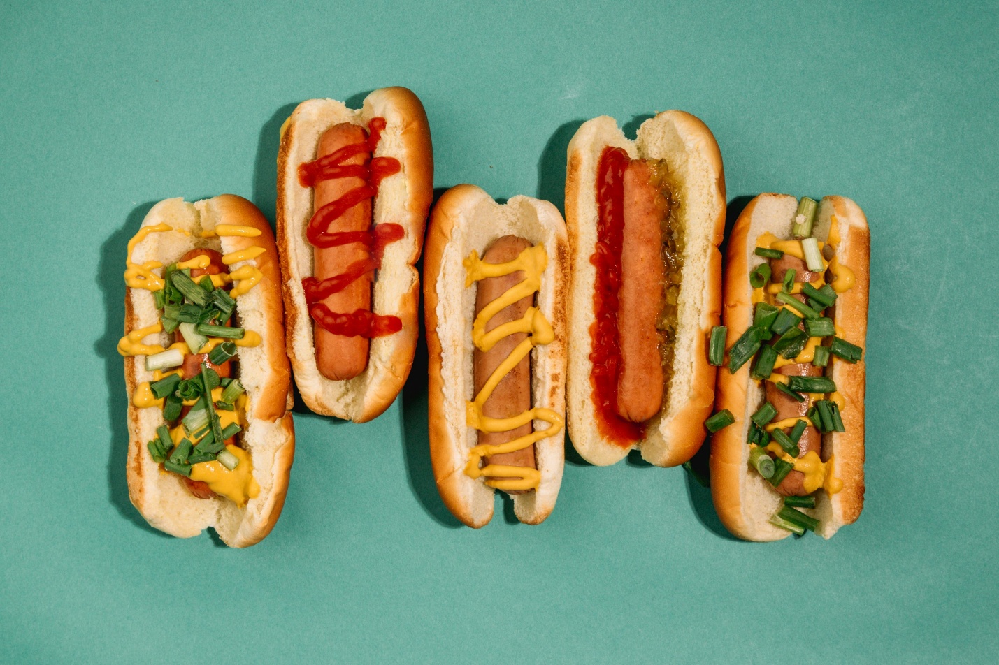 Five colorful hot dogs with various toppings lined up on a green surface