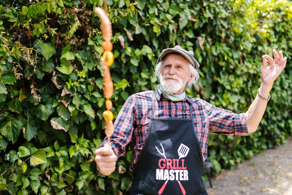 Elderly man wearing a “Grill Master” apron proudly holding a skewer at a community barbecue event.
