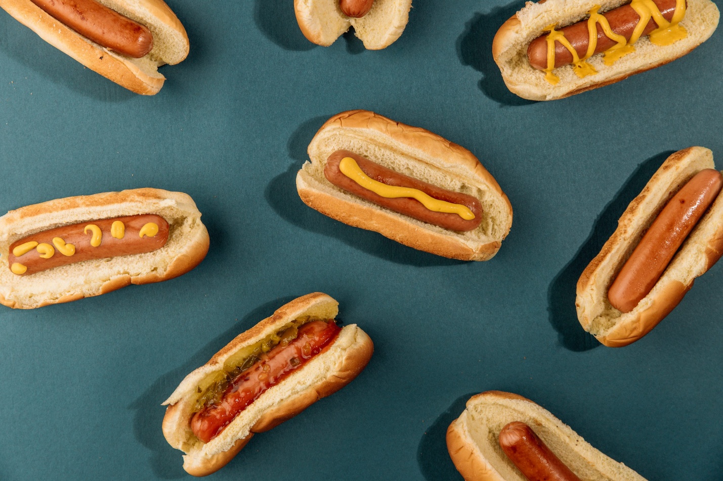 A lineup of hot dogs with mustard and relish on a teal background.