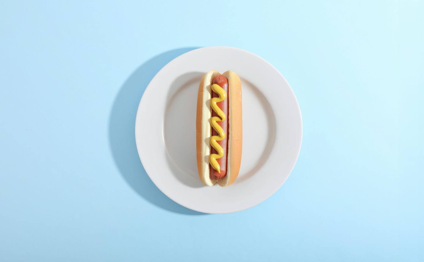 Single hot dog with mustard on a white plate against a pastel blue background